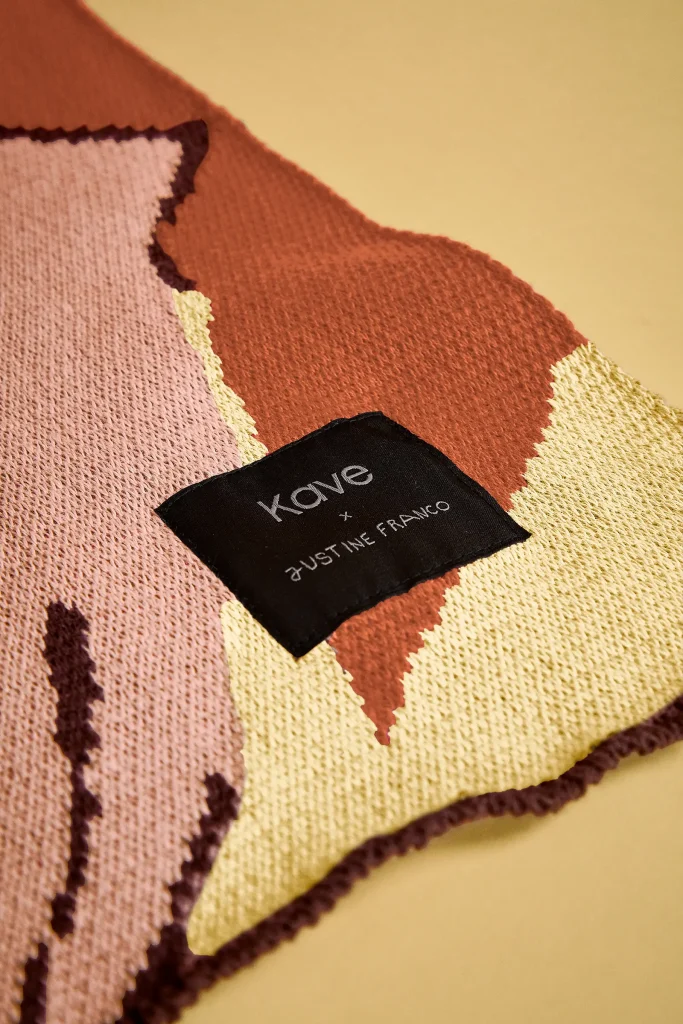 Manta jacquard multicolor de Justine Franco para Kave, con rostros dibujados sobre una butaca de madera.
